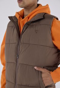 Gilet imbottito marrone con collo alto e chiusura con zip, indossato sopra a una felpa arancione con cappuccio. Presenta un logo triangolare e tasche laterali. Texture liscia.