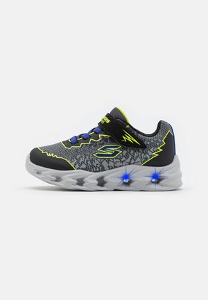 Zapatilla deportiva gris con acentos en negro y amarillo neón, que presenta un upper con patrón, correa ajustable y talones que iluminan. Suela de goma.