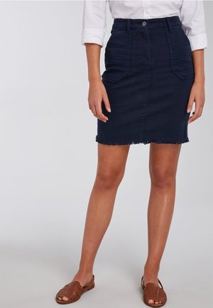 Kobieta ubrana w ciemnoniebieską spódnicę jeansową z przednimi kieszeniami i postrzępionym dołem, białą koszulę z podwiniętymi rękawami oraz brązowe plecione sandały.