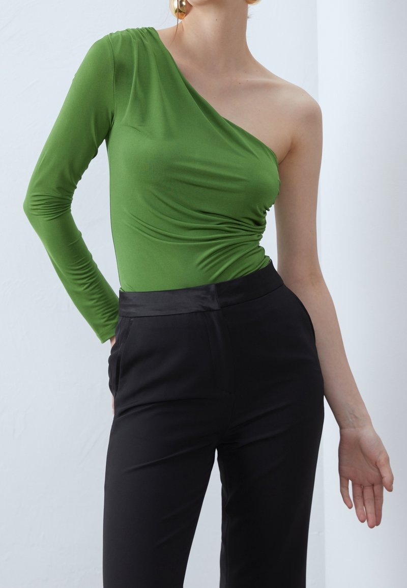 Asymmetrisk grön långärmad topp i elastisk tyg, kombinerad med svarta byxor med en slät midja och fickor.