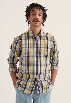 Mann mit lockigem Haar, der ein gelb-grün-navy-kariertes Hemd mit hochgekrempelten Ärmeln und hellblaue Hose trägt, die Hände in den Taschen.