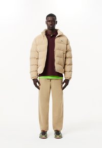 Beige kordflors puffjacka, vinröd polo, grön undertröja, beige byxor och grå sneakers med orangea accenter. Enkel design, avslappnad passform.