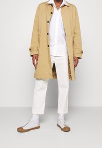 Man in een beige trenchcoat, wit overhemd, witte cropped pants, witte sokken en witte sneakers met bruine zolen.
