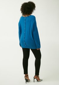 Blauer Strickpullover mit lockerer Passform, der eine Reihe von metallischen Knöpfen hinten hat. Kombiniert mit schwarzen Leggings und silbernen Absätzen.