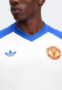 Άνδρας που φοράει λευκή φανέλα του Manchester United με μπλε γιακά και λογότυπο Adidas.