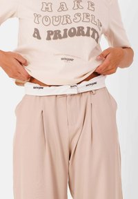 Lichtbeige oversized shirt met tekst; gecombineerd met op maat gemaakte, vloeiende beigebroek en een witte riem met een logo.