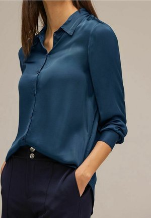 Mujer con una blusa azul oscuro de satén de manga larga metida dentro y pantalones azul marino con una mano en el bolsillo frente a un fondo liso.