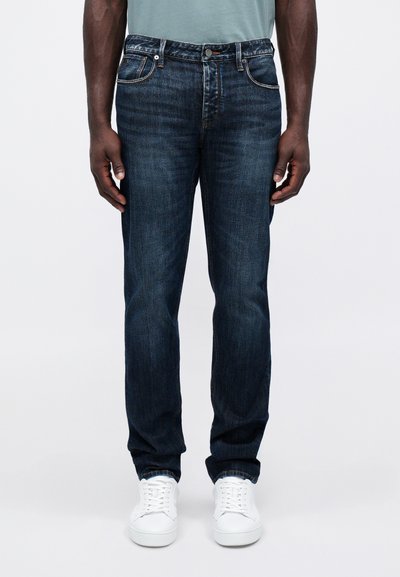 Temno modri jeans s klasičnim krojem, zasnovo s petimi žepi in detajli šivanja. V kombinaciji z belimi supergami za kontrast.