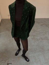 Grön sammetssuitjacka med silvriga nitar, kombinerad med matchande shorts och en svart ribbad polotröja. Svarta skor kompletterar outfiten.