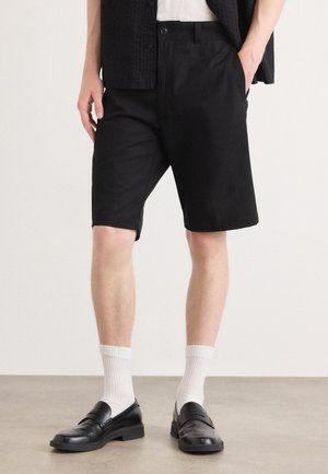Sorte shorts laget av stoff, med en avslappet passform og sidelommer. Brukt med sorte loafers og hvite ankelstrømper.