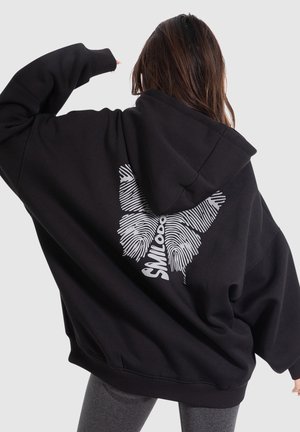 Persoon die een oversized zwarte hoodie draagt met een wit vlinderontwerp in vingerafdrukstijl en de tekst "SMILOOD" op de achterkant.