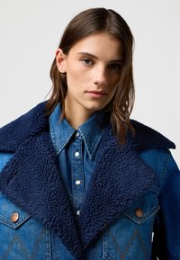 Kurtka dżinsowa z granatowym kołnierzem z imitacji zamszu, wyposażona w duże klapy, jasnoniebieską koszulę dżinsową pod spodem oraz brązowe guziki.