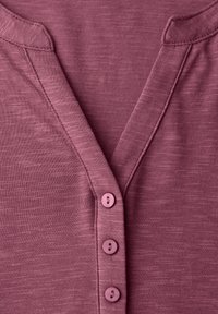 Zbliżenie na wrzosowy dzianinowy sweter z dekoltem w serek i trzema dopasowanymi guzikami z przodu.