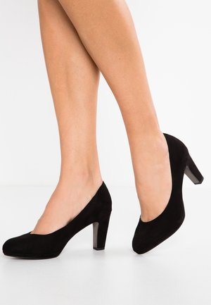 Woman wearing classic black suede mid-heel pumps, standing with one foot slightly raised, against a plain white background.