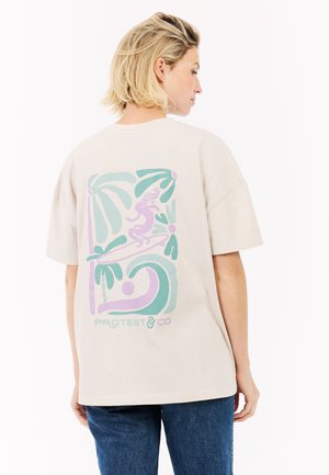 Vrouw met kort blond haar draagt een beige oversized T-shirt met pastel tropische afbeelding en blauwe jeans, staand tegen een witte achtergrond.