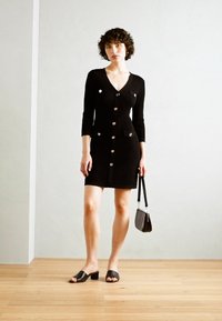 Robe noire en tricot avec un col en V, des boutons et des poches avant. Le mannequin tient un petit sac et porte des sandales noires �à glissière. Sol en bois clair.