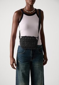 Persoon in een mouwloos wit topje met zwarte randen, donkere wijde jeans, zilveren ketting en een zwart crossbody geborduurde tas.