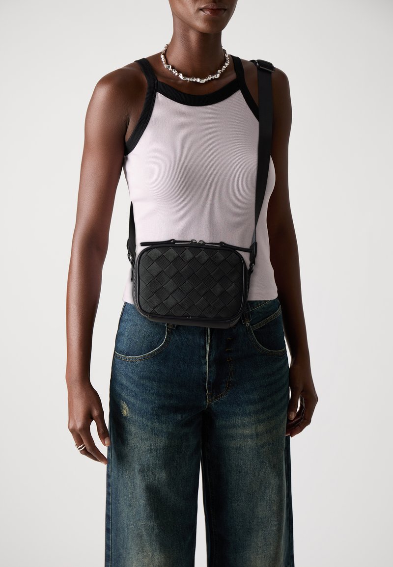 Persoon in een mouwloos wit topje met zwarte randen, donkere wijde jeans, zilveren ketting en een zwart crossbody geborduurde tas.