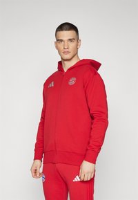 Felpa rossa con zip realizzata in tessuto morbido, dotata di cappuccio, tre strisce bianche sulla spalla sinistra e un logo circolare sul petto.