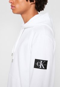 Felpa bianca con cappuccio e tasca frontale, caratterizzata da un'etichetta nera con la scritta "Calvin Klein Jeans" sulla manica sinistra e dettagli in metallo argentato.