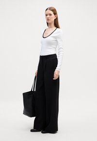 Fehér, hosszú ujjú felső fekete, kerek nyakkivágással, magas derekú fekete, bő szárú nadrággal párosítva. A modell egy fekete kézitáskát tart.