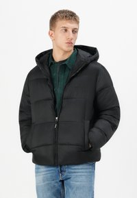 Sort puffjakke med hætte, der har en quiltet design, lynlås og sidelommer, båret over en grøn skjorte.