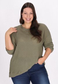 Olivgrön, strukturerad tröja med rund halsringning, långa ärmar och asymmetrisk fåll, kombinerad med mörkblå jeans.