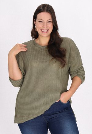 Pulover texturat de culoare verde măsliniu, cu guler rotund, mâneci lungi și tiv asimetric, asortat cu blugi de culoare albastru închis.