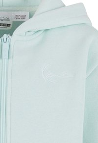 Šviesiai mėlyna zip-up džemperis, pagamintas iš minkštos medžiagos, su mažuoju baltu siuvinėtu logotipu ir kapuci. Priežiūros etiketė matoma viduje.