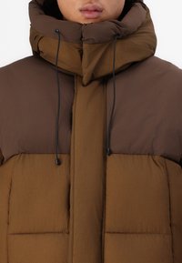 Braune Pufferjacke mit hohem Kragen und verstellbarem Kapuzen. Sie verfügt über eine gesteppte Textur, kontrastierende Brauntöne und schwarze Kordelakzente.