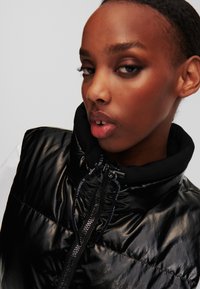 Vest puf negru cu textură lucioasă și netedă, guler înalt și detalii cu fermoar. Prezintă un logo îndrăzneț pe toată lungimea fermoarului și un design plin, matlasat.