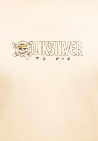 Κρανίο που φοράει ψάθινο καπέλο με σταυρωμένα οστά πίσω, δίπλα σε στυλιζαρισμένο κείμενο "Quiksilver" και ιαπωνικούς χαρακτήρες σε κρεμ ύφασμα.