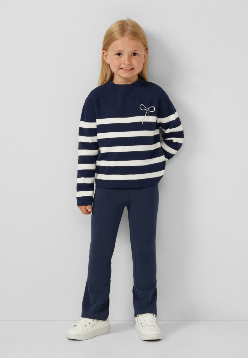 Maglione a righe blu navy con strisce bianche, presenta un fiocco decorativo, abbinato a leggings blu navy e sneaker bianche, texture liscia.