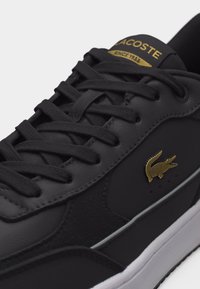Μαύρο αθλητικό παπούτσι Lacoste με χρυσλό λογότυπο κροκοδείλου, μαύρα κορδόνια και λευκή σόλα, που δείχνει κοντινή λήψη των πλευρικών και επάνω λεπτομερειών.