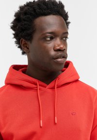 Młody mężczyzna z krótkimi, kręconymi włosami, w jasnoczerwonym hoodie, patrzy w bok na tle jednolitego, białego tła.