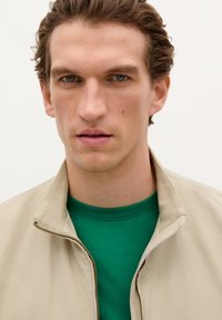 Chaqueta cremosa con cremallera y cuello alzado sobre una camiseta verde. Textura suave y diseño ajustado con un brillo sutil en la chaqueta.
