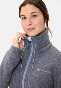 Giacca in pile grigia con colletto alto, dotata di zip frontale e cordini. Logo sul petto e dettagli in tessuto testurizzato.