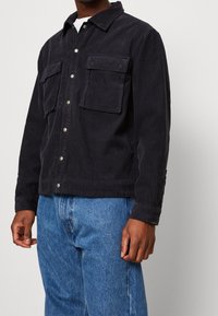 Navy modrá kordurová bunda se dvěma předními kapsami, zapínáním na knoflíky a límečkem. V kombinaci s modrými džíny.