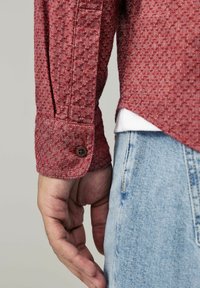 Chemise rouge texturée avec des poignets à bouton, affichant un motif en losanges. Portée sur un jean en denim bleu clair avec un léger effilochage à l'ourlet.