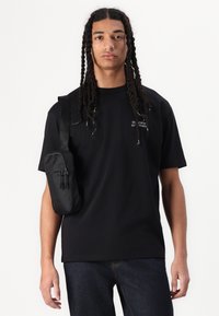 Sort t-skjorte med kort erm og logo-detalj, kombinert med mørk denim jeans. En svart poseveske bæres over skulderen.