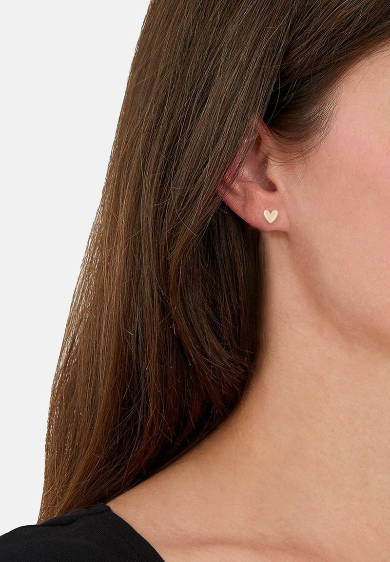 Persona con cabello castaño que lleva un pequeño pendiente en forma de corazón beige en la oreja derecha, visible contra un fondo claro y con una camiseta negra.