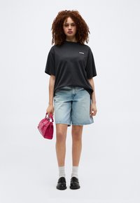 Γυναίκα με σγουρά κόκκινα μαλλιά που φοράει μια μαύρη φαρδιά μπλούζα Levi's, ανοιχτό μπλε τζιν σορτς, μαύρα μοκασίνια, άσπρες κάλτσες, κρατώντας μια μικρή ροζ τσάντα.