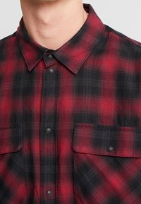 Röd och svart rutig skjorta med knäppning i kragen, två framlommor och svarta tryckknappar. Mjuk, texturerad tyg med fiskbensmönster.