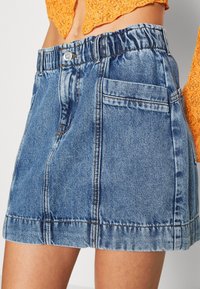 Jeanskjol med elastisk midja, ljusblå färg, som har två framfickor och kontrasterande sömmar. Kort, A-linjeformad design.