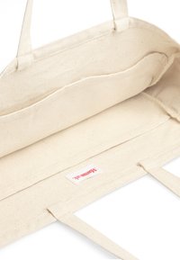 Beige bomullsaxelväska med två handtag, öppen topp och en sydd etikett. Interiören har en enkel, omönstrad textur.