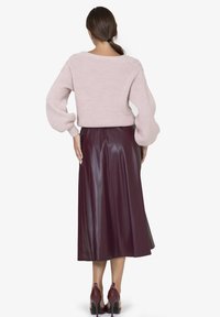 Frau mit braunem Haar im Pferdeschwanz, die einen rosa gestrickten Pullover, einen burgunderfarbenen Leder-Midirock und passende High Heels trägt und von hinten schaut.