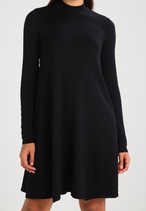 Femme portant une robe noire à manches longues, longueur genou, coupe trapèze avec un col montant, sur un fond uni.