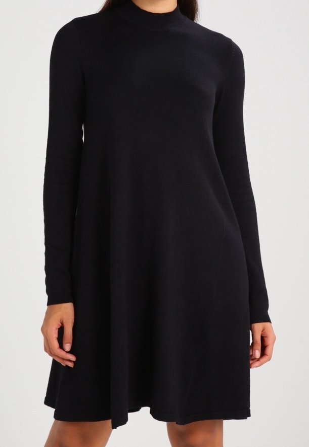 Femme portant une robe noire à manches longues, longueur genou, coupe trapèze avec un col montant, sur un fond uni.