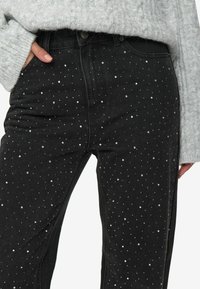 Schwarze Jeans aus Denim mit verstreuten silbernen Nieten, hoch tailliertes Design, mit einem teilweise sichtbaren strukturierten, grauen Strickpullover.