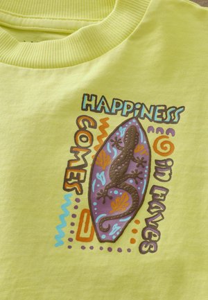 Gul bomuld t-shirt med et gekko-print og teksten "LYKKE KOMMER I BØLGER" i brune nuancer og farverige accenter. Glat tekstur.